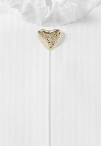 Tela blanca con rayas verticales presenta un cuello con volantes y un brillante cierre en forma de corazón dorado en el centro, añadiendo un toque decorativo.