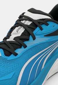Scarpa sportiva blu con accenti neri, realizzata in materiale a rete, strisce bianche e lacci neri. Forma curva e dettaglio della superficie texturizzata.