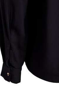 Poignet de chemise noire à manches longues avec fermeture par bouton, montrant la texture du tissu et les détails de couture sur la manche et l'ourlet.