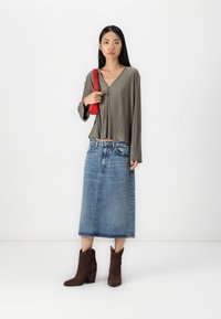 Olivgrüne, plissierte Bluse mit langen Ärmeln und V-Ausschnitt, kombiniert mit einem blauen Denim-Midirock und braunen Wildleder-Stiefeletten. Rote Schultertasche.