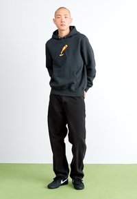 Sort hættetrøje med et orange og gult grafisk design, parret med sorte løse jeans og sorte sneakers. Glat materiale, afslappet pasform.