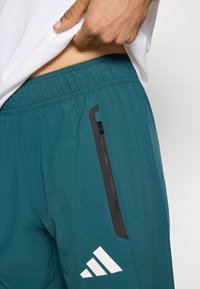 Türkiz atlétikai rövidnadrágok könnyű anyagból, rugalmas derékpánttal és cipzáras zsebbel fekete részletekkel. Fehér logó látható.