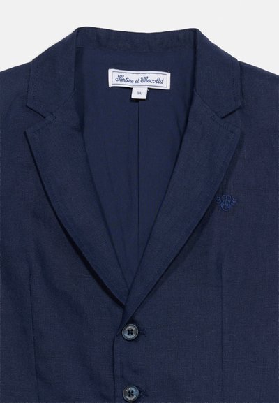 Μπλέ σκούρο μπλέιζερ παιδικό με πέτα, δύο κουμπιά, κεντημένο λογότυπο στο στήθος και λευκή ετικέτα επωνυμίας "Tartine et Chocolat" μέσα στη γιακά.