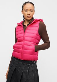 Νεαρή γυναίκα με πλεξούδες που φοράει φωτεινό ροζ γεμιστό γιλέκο πάνω από καφέ μακρυμάνικο ριμπ πουκάμισο και σκούρα πλισέ φούστα.