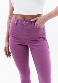 Jeans de cintura alta, ajustados, en un vibrante denim morado, con bolsillos frontales y cierre de botón, que presentan una textura suave y un ligero estiramiento.