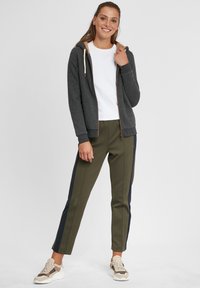 Haut à capuche zippé gris foncé avec doublure beige, associé à un pantalon vert olive avec des rayures latérales bleu marine et des baskets claires. Look athlétique décontracté.