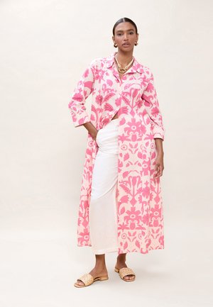 Mujer vistiendo un abrigo largo con estampado floral en rosa y blanco, pantalones blancos, sandalias beige tejidas y joyería dorada, de pie con una mano en el bolsillo.