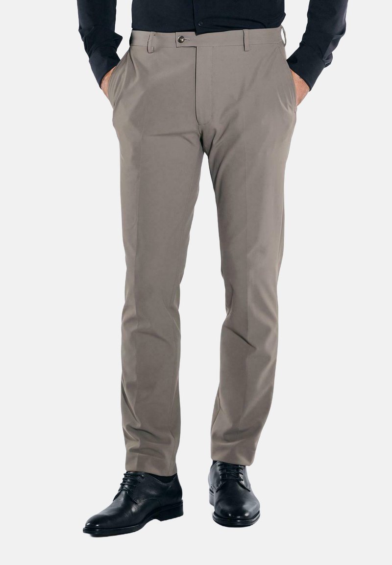Mann trägt beige Anzughose, schwarze Anzugschuhe und ein dunkles langärmeliges Hemd, steht mit den Händen in den Taschen.