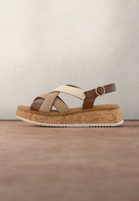 Sandale plateforme avec des brides croisées beige et marron et une semelle à texture liège avec une boucle argentée sur la bride de cheville.