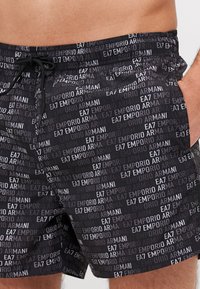 Fekete fürdősort, amelyen ismétlődő logóminta található különböző szürke árnyalatokban, rugalmas derékkal és állítható zsinórral.