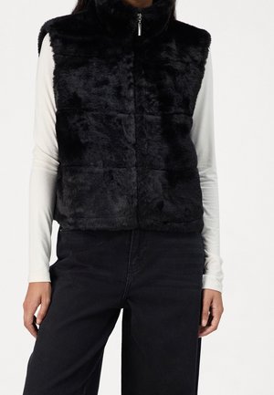 Persoon draagt een zwart, donzig mouwloos vest met een rits aan de voorkant over een wit shirt met lange mouwen en zwarte broek, staand tegen een witte achtergrond.