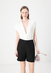 Hvit kortermet bodysuit med dyp V-utringning og glidelås, parret med høyveide svarte shortser. Modellen holder en liten rosa veske.