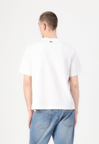 Camiseta blanca de algodón con mangas cortas, cuello redondo y pequeño logo de cocodrilo verde en la parte superior de la espalda; combinada con jeans de denim azul.