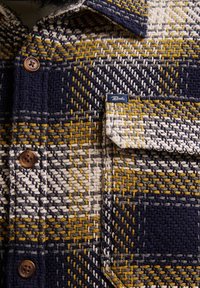 Camicia con bottoni in tessuto testurizzato, caratterizzata da un motivo a quadri blu navy, bianco e senape. Include una tasca sul petto e bottoni marroni.