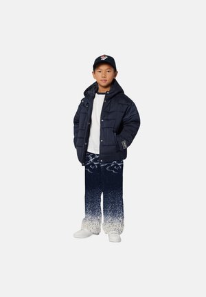 Marineblauer Pufferjacke mit Kapuze, weißem T-Shirt, gradient blau-weiß gemusterten Hosen und weißen Sneakers. Die Mütze hat ein grafisches Design.