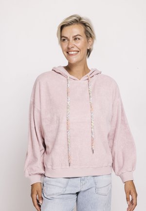 Smiling woman with short blonde hair wearing a pastel pink textured hoodie with colorful drawstrings and light blue jeans, standing against white background.