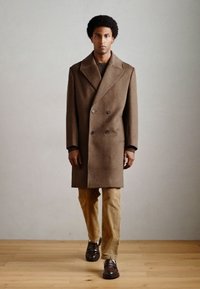 Cappotto doppio petto marrone con collo a lancia, abbinato a pantaloni di corduroy beige strutturati e mocassini marrone scuro, indossato da un modello su uno sfondo chiaro.