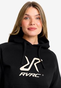 Svart huvtröja tillverkad av texturerat tyg med en stor, krämfärgad RVRC-logotyp på framsidan, med dragsko i huvan och avslappnad passform.