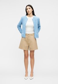 Γυναίκα που στέκεται με τα χέρια πίσω, φοράει ανοιχτό μπλε υφασμάτινο σακάκι, λευκό τοπ, μπεζ σορτς και λευκές μυτερές γόβες.