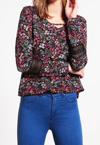 Květinová blůza na černém pozadí s růžovými a bílými květy, strukturovanými rukávy a peplum pasem, doplněná modrými džínami.