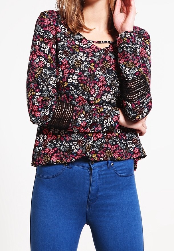 Květinová blůza na černém pozadí s růžovými a bílými květy, strukturovanými rukávy a peplum pasem, doplněná modrými džínami.