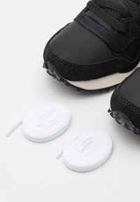 Chaussures de sport noires avec une tige en nylon lisse, des accents en daim et une semelle en caoutchouc texturée. Deux lacets blancs roulés sont à proximité.