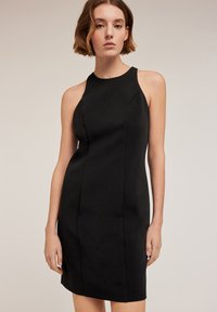 Svart ärmlös klänning med en figursydd siluett, mjuk textur och detaljer i framsömmen. Längden går till mitten av låret och har en rundad halsringning.