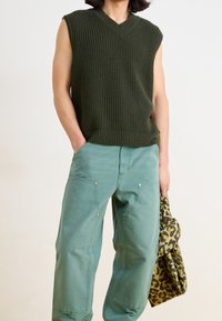 Ärmlös, mörkgrön stickad tröja med V-ringning; kombinerad med ljusgröna, löst sittande byxor med synliga fickor och subtila gulddetaljer. Håller en leopardmönstrad väska.