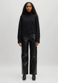 Schwarzer Rollkragenpullover mit geripptem Saum, kombiniert mit glänzenden, schwarzen Hose im Krokodilmuster mit weitem Bein und schwarzen Stiefeletten.