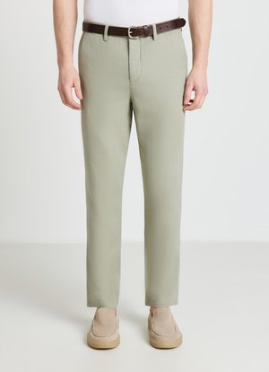 Homme portant un pantalon droit vert clair avec une ceinture marron et des chaussures beige sans lacets, debout devant un fond blanc.
