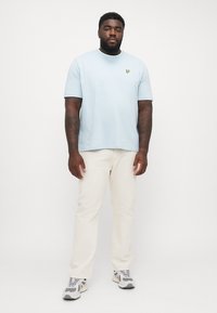 Lys blå bomulds t-shirt med rund halsudskæring og lille logo på brystet, parret med beige bukser og grå sneakers.