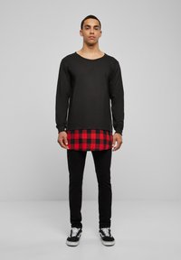 Chemise noire à manches longues portée sur un tissu à carreaux rouges et noirs, associée à un jean skinny noir et des baskets noires avec des accents blancs.