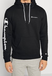 Haut à capuche noir en mélange de coton, avec un col zippé, des cordons blancs, une poche avant et le logo "Champion" en blanc sur la manche et la poitrine.
