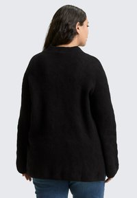 Vrouw met lang donker haar, gekleed in een zwarte, gestructureerde trui en een blauwe spijkerbroek, gezien van achteren tegen een lichtgrijze achtergrond.