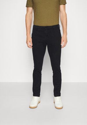 Hombre con pantalones negros ajustados, camiseta verde oliva y zapatillas blancas, de pie contra un fondo claro y liso.