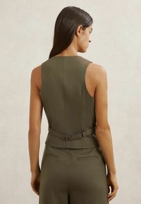 Donna vista di spalle che indossa un gilet senza maniche verde oliva con fibbia regolabile sul retro e pantaloni abbinati a vita alta.
