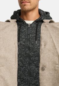 Cappotto beige in misto lana con collo e dettaglio a bottoni, indossato sopra una felpa grigia con zip caratterizzata da un motivo strutturato e cappuccio con coulisse.