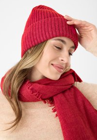 Rode geribbelde gebreide muts met een bijpassende rode fringesjaal. Beide zijn gemaakt van zachte, gezellige materialen, die textuur en warmte aan de outfit toevoegen.