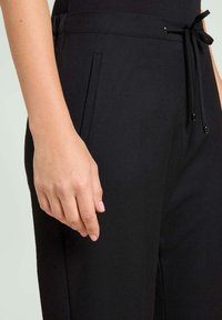 Pantalon noir en tissu doux, doté d'une taille à cordon, de poches latérales et d'une coupe droite élégante. Main posée près de la taille.