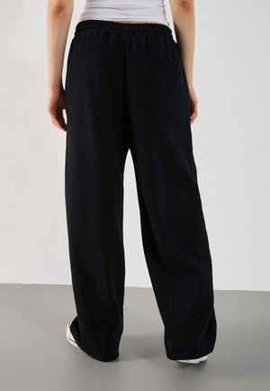 Personne debout, portant un pantalon noir ample avec une taille élastique et une poche arrière, des baskets blanches et un top blanc sans manches.