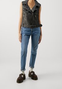Ärmlös svart läderväst med bälte i midjan, kombinerad med ljusblå, figursydda jeans och bruna skor med utskärningar.