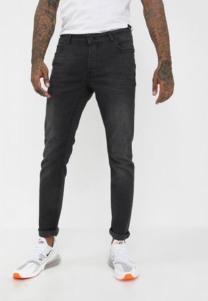 Homme portant un jean noir slim-fit avec les revers roulés et des baskets blanches Nike Air Max 270, debout les mains détendues le long du corps.