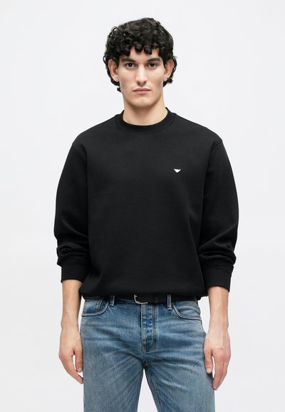 Čierna mikina s dlhými rukávmi a okrúhlym výstrihom, s malým bielym logom na hrudi. Nosená s svetlomodrými džínsami.