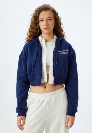 Cropped navy hoodie in soft material with zip closure, front pockets, and white text branding. Paired with light-colored elastic waist pants.