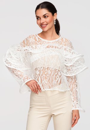 Femme aux cheveux foncés attachés en queue de cheval portant un chemisier blanc en dentelle à manches longues à volants et un pantalon crème, souriante et posant avec la main sur la hanche.