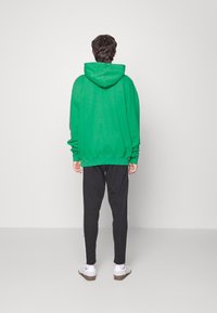 Una persona sta in piedi con le spalle alla fotocamera, indossando un hoodie verde, pantaloni neri con strisce bianche e sneakers bianche su uno sfondo chiaro.