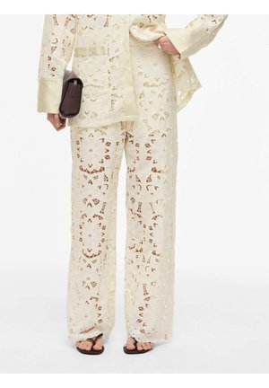 Gebroken wit gebloemde kanten broek en bijpassende jas met brede manchetten, gecombineerd met zwarte sandalen en een kleine donkere clutchtas.