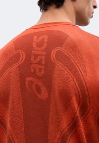 Άτομο που φοράει πορτοκαλί μπλουζάκι Asics, δείχνοντας την πλάτη με το όνομα της μάρκας και το λογότυπο υφασμένα στο ύφασμα.