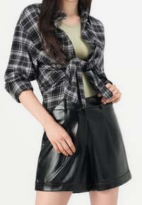 Svart och grå rutig skjorta med knappar, knuten i midjan, lager på en ljusgrön åtsittande topp, tillsammans med högmidjade glansiga svarta shorts.