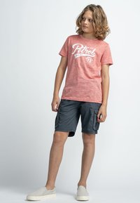 Punainen marmoroitu T-paita, jossa on valkoinen "Petrol" -graafika, yhdistettynä harmaisiin cargo-shortseihin, joissa on sivutaskut, ja vaaleanharmaisiin liukuvaan kenkään.